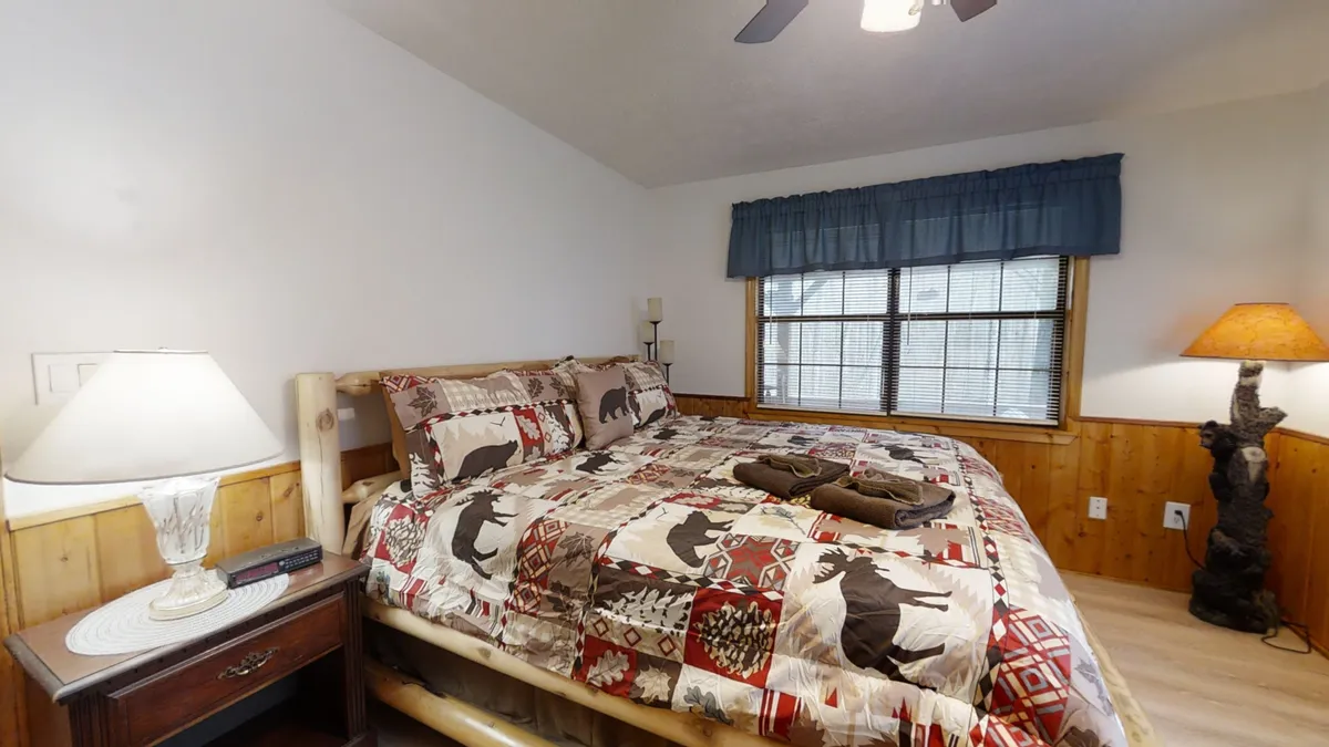 A king-sized log bed with a colorful quilt sits next to a window with drapes, accompanied by a nightstand and a soft decorative lamp. The warm, comforting atmosphere invites couples to relax, share stories, and enjoy a quiet morning coffee, reinforcing the sense of privacy and retreat.