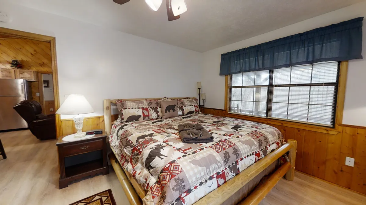 A spacious bedroom featuring a large bed with a patterned quilt, inviting wooden furnishings, and a window with blinds that offers a view out to the natural surroundings. The soft lighting and warm decor evoke comfort after active days.