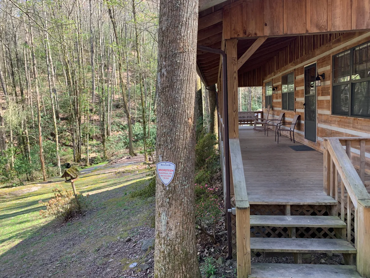 A welcoming wooden porch extends from the cabin, surrounded by tall trees and lush greenery. The porch offers comfortable seating ideal for relaxing, sharing meals, or simply soaking in the sunrise and evening views. The space is perfect for family gatherings or quiet moments enjoying the natural setting.
