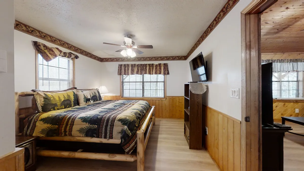 A cozy bedroom featuring a bed with patterned bedding, sturdy wooden furniture, and large windows that allow natural morning light to filter in. The scene evokes comfort and rustic charm, making it easy to imagine a couple waking up to the sounds of birds and a babbling creek just outside.