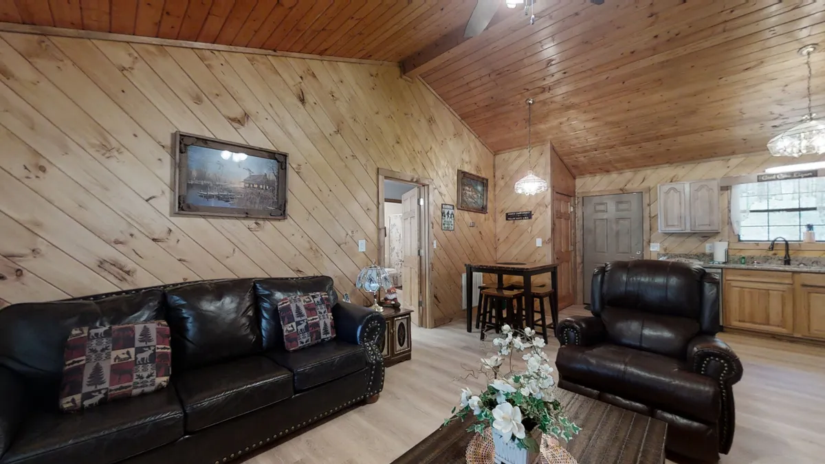 This image features an inviting living room with leather sofas, a wooden ceiling, a dining table, and decorative wall art. The room is warm and spacious, designed for relaxation, entertaining friends, or family gatherings. The layout shows the seamless flow between living and dining spaces, providing ample room for everyone to connect and unwind.