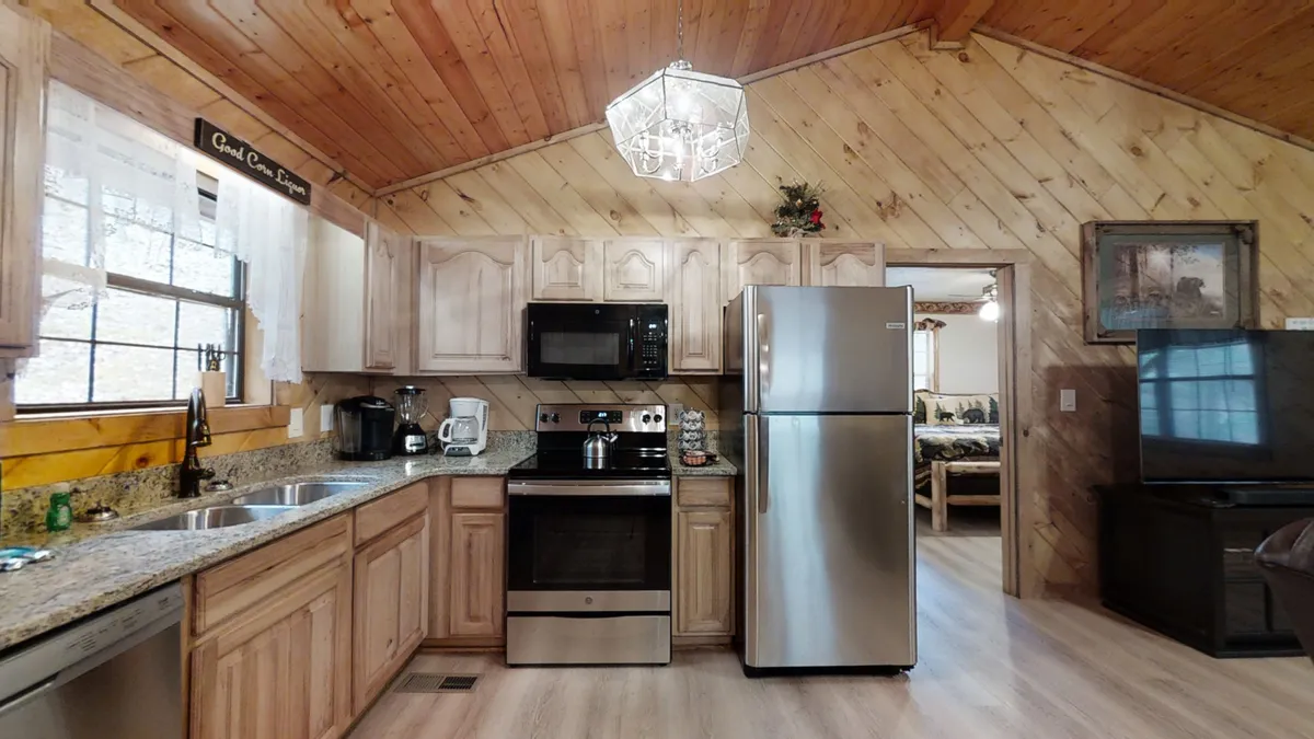 The image presents a sunlit kitchen with granite countertops, stainless steel appliances, wooden cabinetry, and a window allowing natural light to flow in. The layout hints at easy movement between kitchen and dining areas, reinforcing a sense of openness.