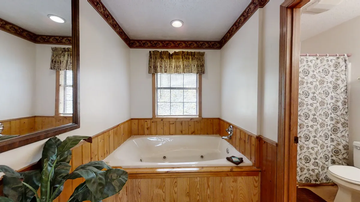A tranquil bathroom scene featuring a spacious jacuzzi tub beneath a curtained window. Wood paneling and a large mirror complete the relaxing setup, perfect for unwinding in privacy.